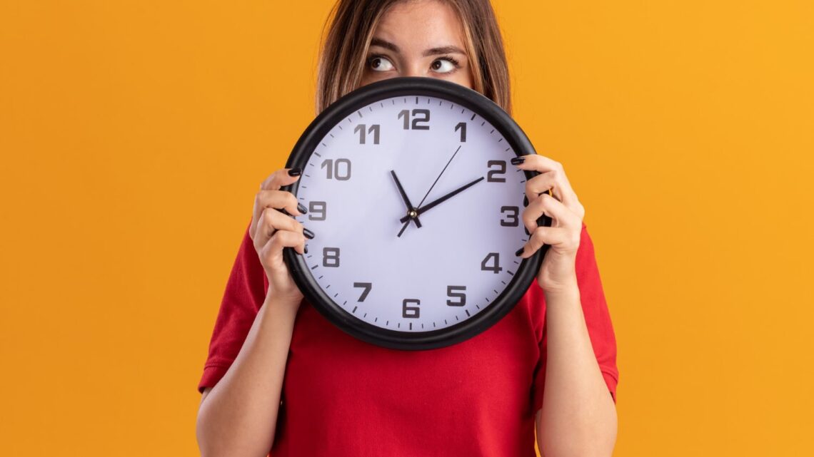 confused-young-pretty-woman-holds-clock-looking-side-isolated-orange-wall-min